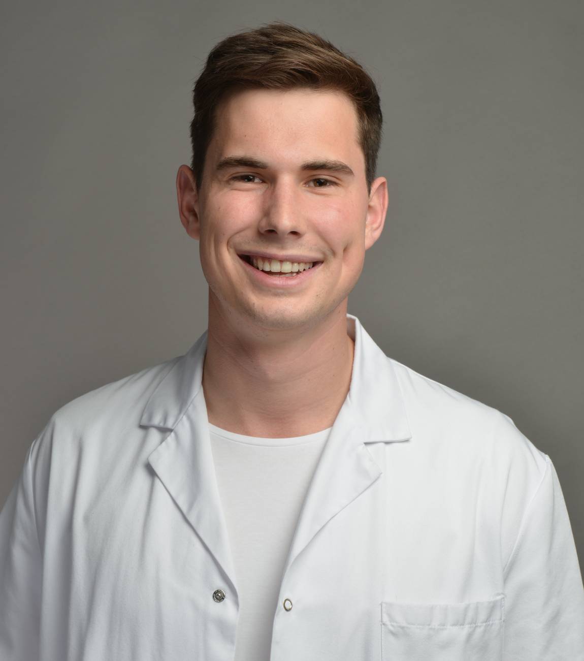 Ein junger Mann mit kurzen braunen Haaren lächelt in die Kamera und trägt einen weißen Laborkittel über einem weißen Hemd. Der Hintergrund ist einfarbig grau.
